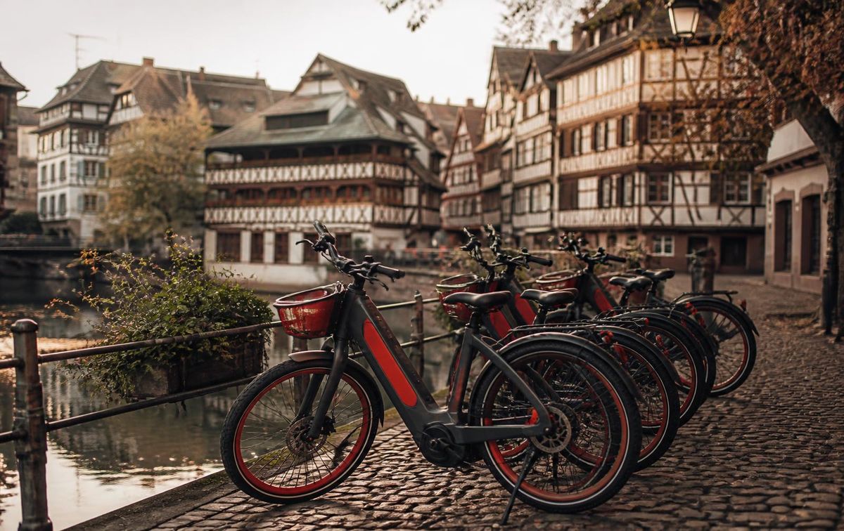 A row of red-accented electric city bikes parked on a cobbled quay in Petite France, Strasbourg, with half-timbered houses behind