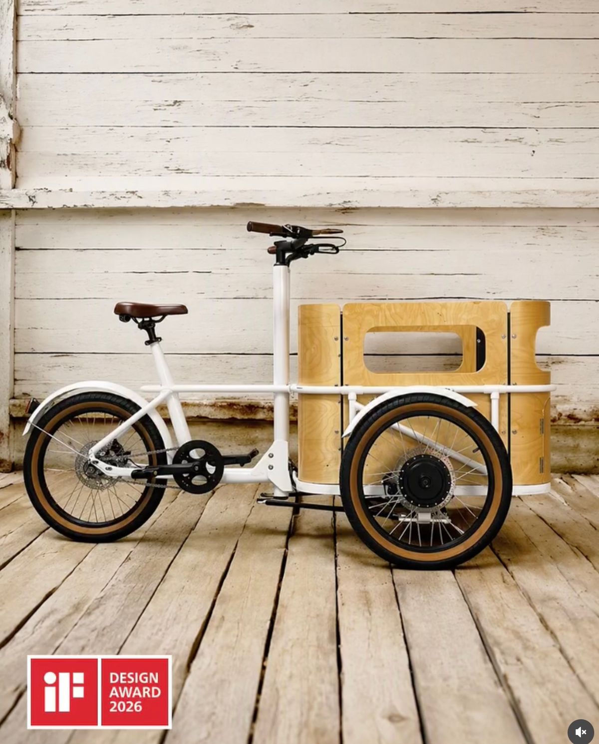 The white minica electric cargo trike with its birch plywood box, winner of the iF Design Award 2026, photographed against a weathered white timber wall