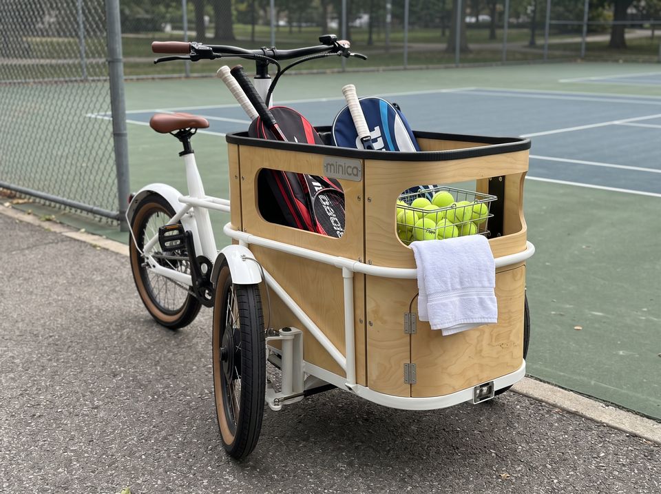 A minica electric cargo trike loaded with racket bags and a basket of tennis balls beside a court.